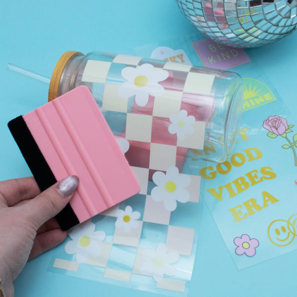 Pink sanding tool being used on a checkered design on a clear container with flowers, on a blue background with text and stickers.