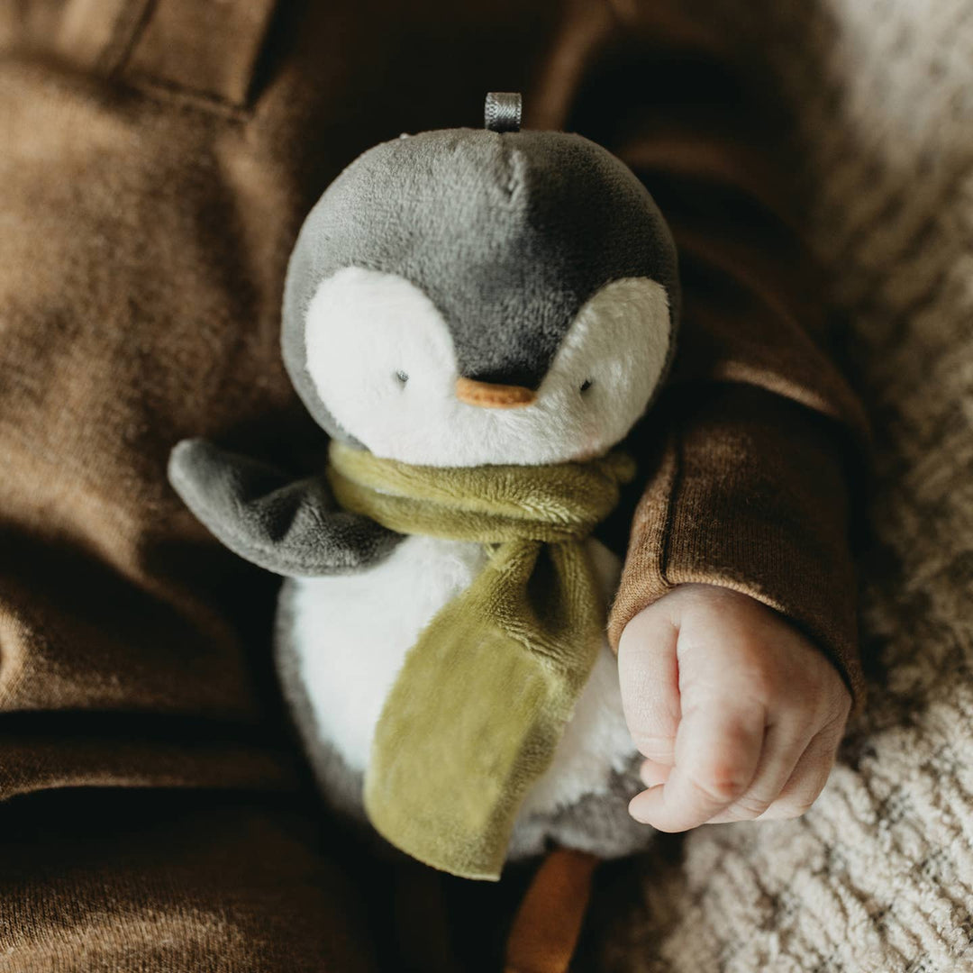 Child's hand holding a plush penguin toy with a green scarf on a soft surface.