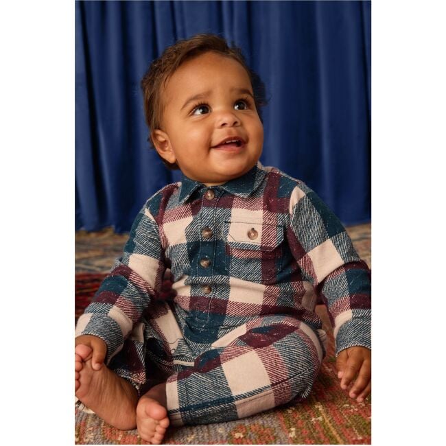 Baby wearing a plaid outfit sitting on a patterned rug with a blue curtain background