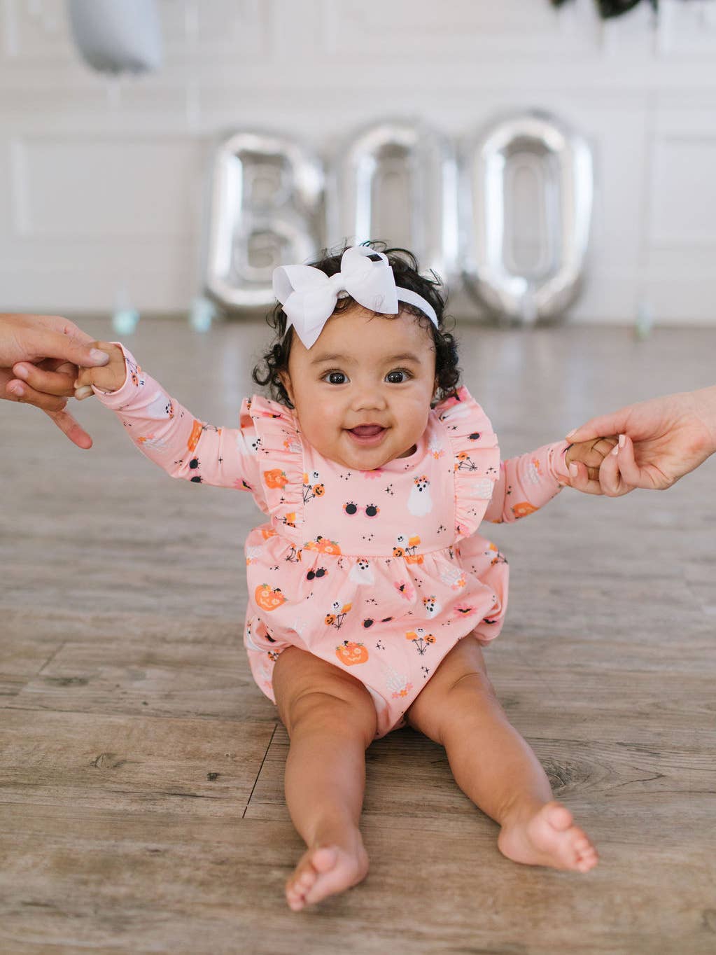 Baby in a pink onesie with Halloween-themed print sitting on a wooden floor, holding hands.