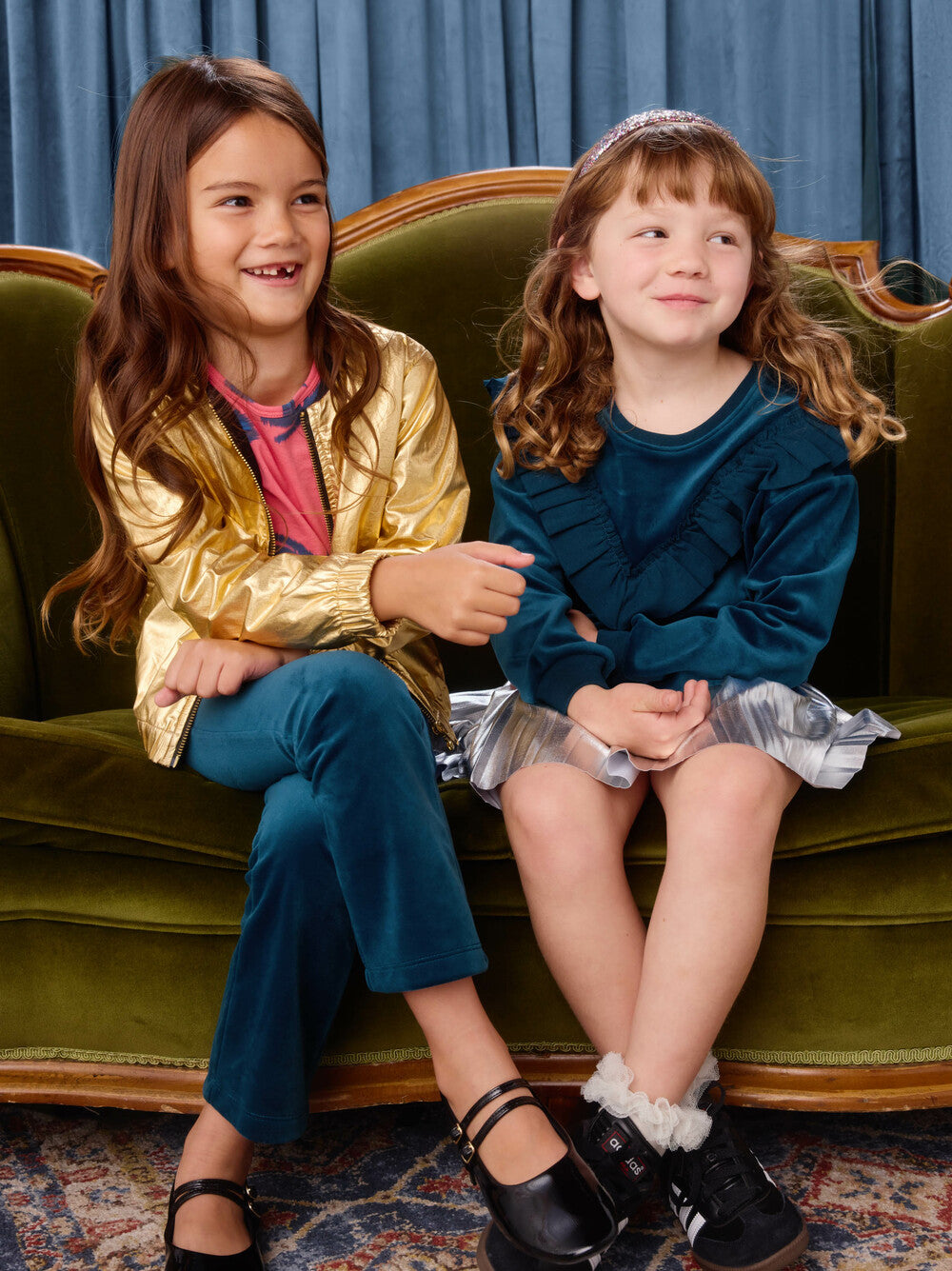 Two young girls sitting on a green couch wearing colorful outfits.