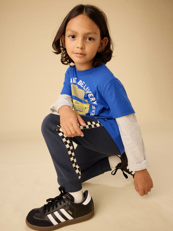Child wearing a blue shirt with text, white long-sleeve shirt, dark pants, and black Adidas shoes on a beige background