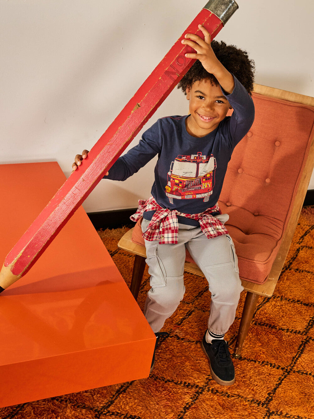 Child holding a large red pencil in a room with orange furniture and a patterned rug.