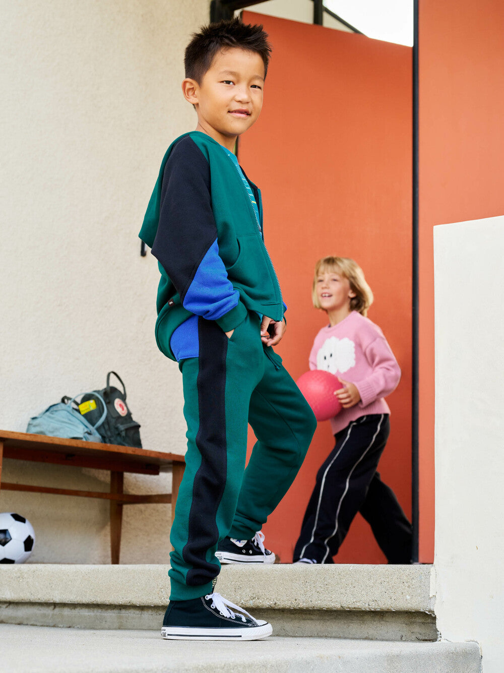 Child in jade green and navy tracksuit standing on steps with another child in pink shirt in the background.