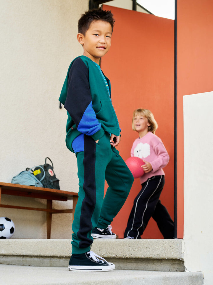 Child in jade green and navy tracksuit standing on steps with another child in pink shirt in the background.
