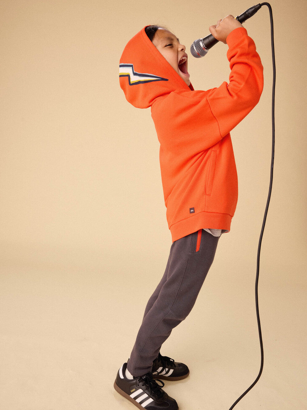 Person wearing an orange hoodie and gray pants, holding a microphone on a beige background