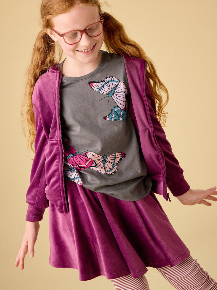 Young girl wearing a gray shirt with butterfly designs and a purple jacket on a beige background