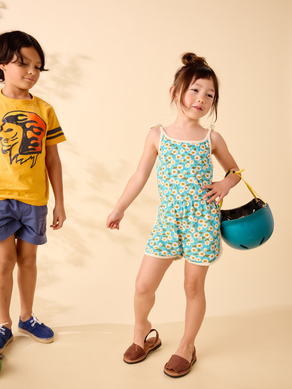 Two children, a boy and a girl, standing against a beige background. The boy is wearing a yellow shirt with a lion design and blue shorts, while the girl is in a floral romper holding a blue helmet.