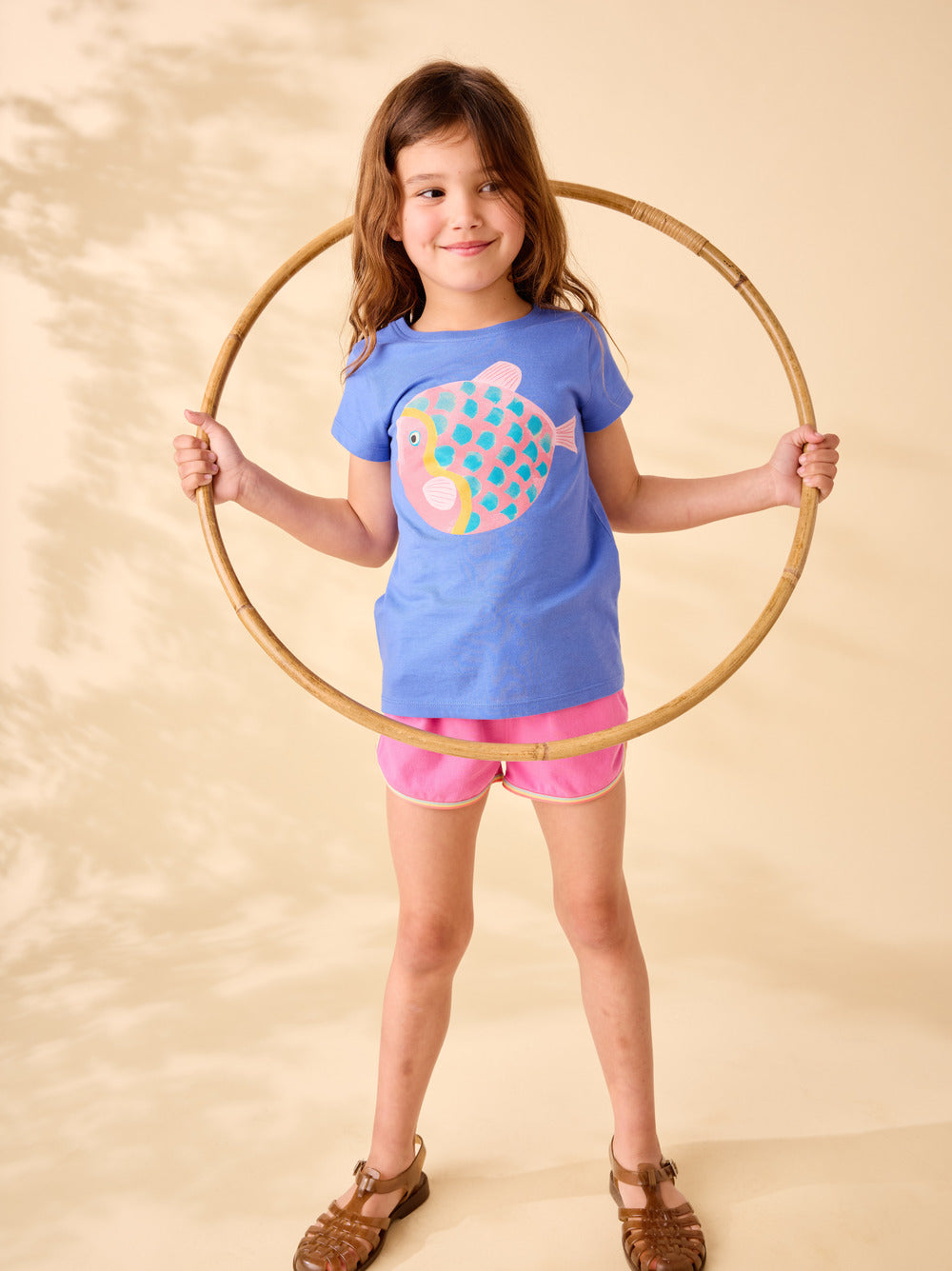 Young girl holding a hula hoop with a blue shirt featuring a colorful fish design on a beige background
