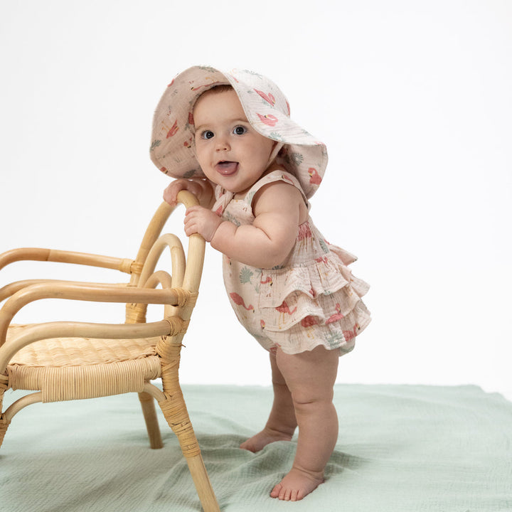 Baby in a floral outfit and hat standing next to a chair on a light green blanket.