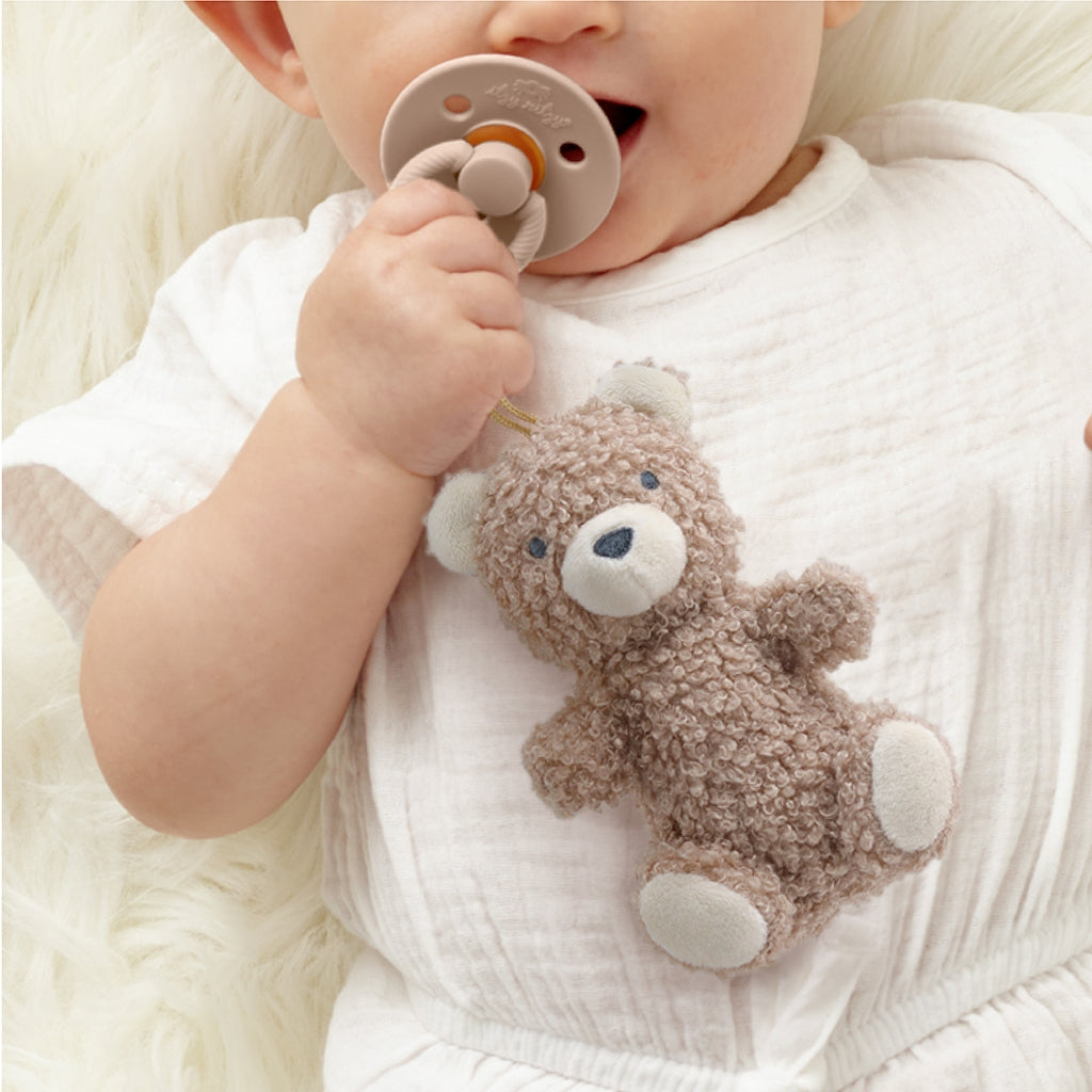 Baby holding a teddy bear rattle and a pacifier on a soft surface