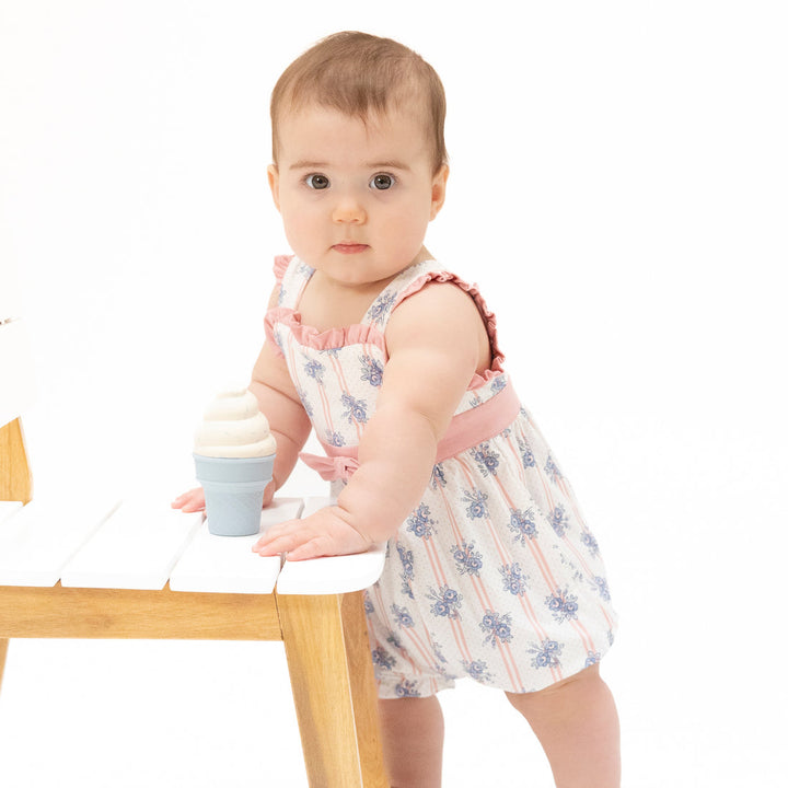 Baby in a floral dress standing next to a small table with a cupcake on a white background