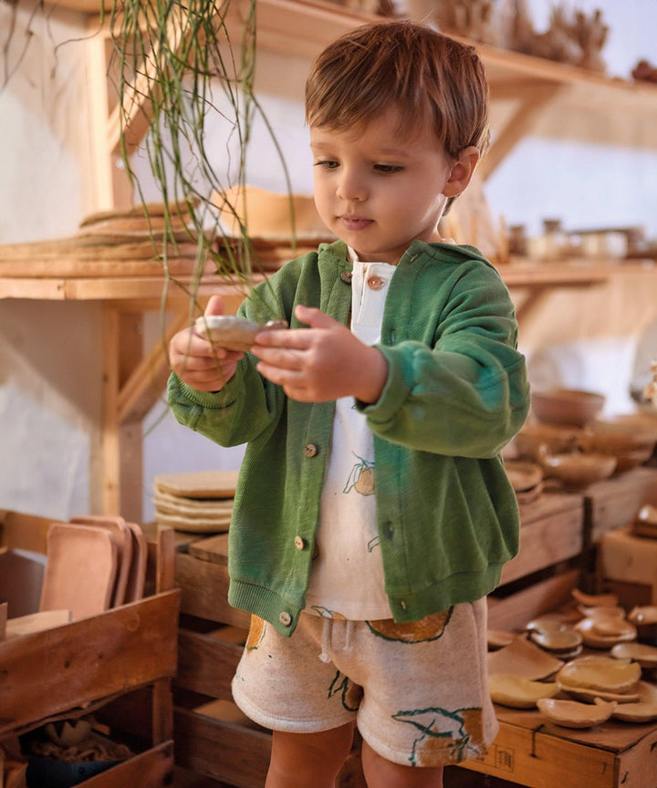 young boy modeling the heathered beige shorts with big yellow oranges on the shorts with drawstring and a green sweatshirt jacket. 