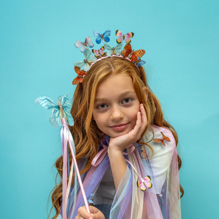 Young girl wearing a butterfly-themed headband and wand against a blue background