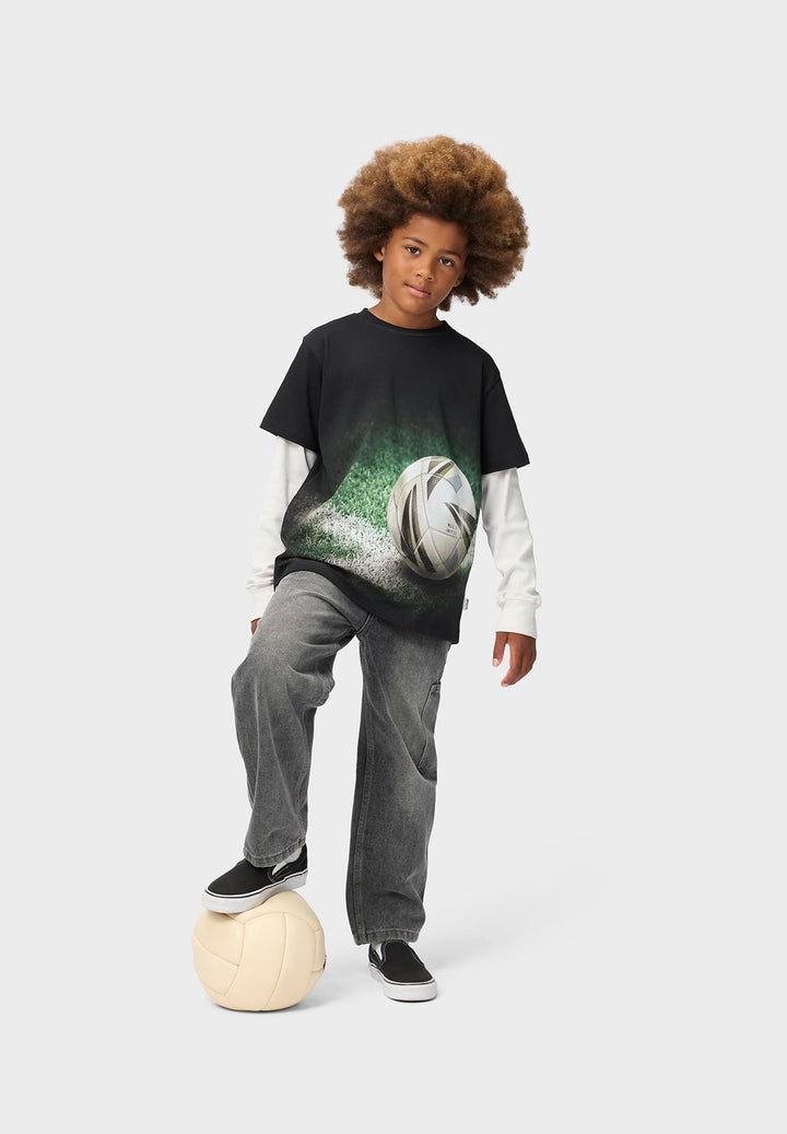 Child wearing a black t-shirt with a graphic design, standing on a soccer ball against a white background