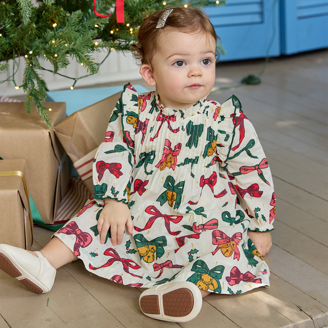 Child wearing a festive dress with bow pattern sitting on the floor near Christmas decorations.