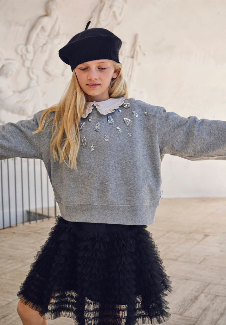 Young girl wearing a gray sweater with silver tear drop embellishments and a black lace skirt, standing indoors.