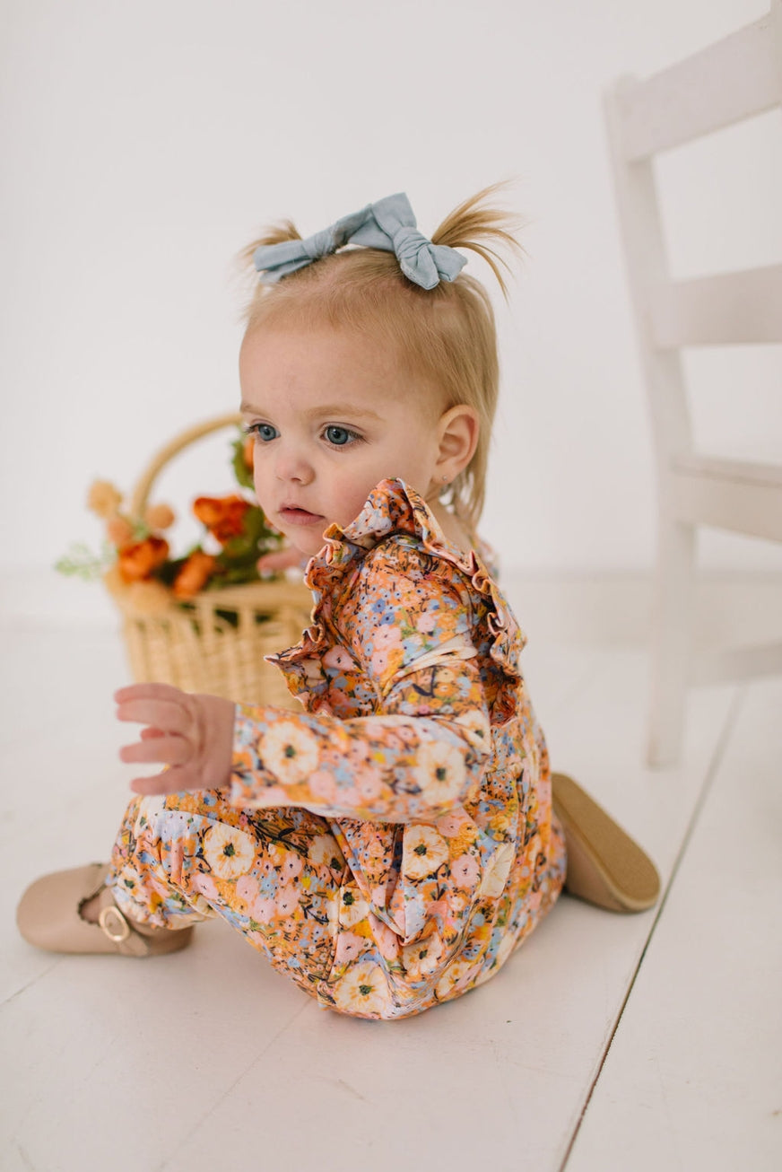 Baby in a floral outfit sitting on a white floor with a basket of flowers.