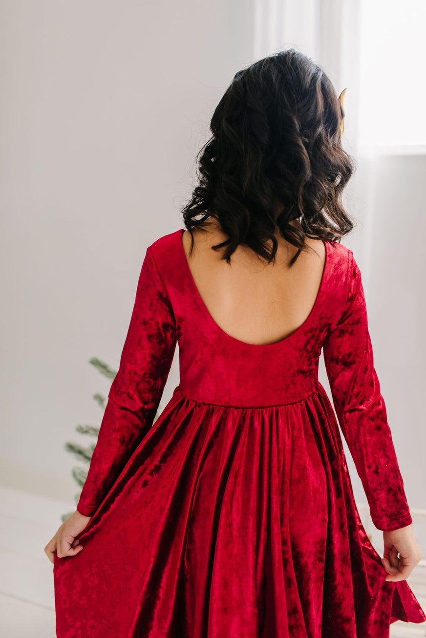 child wearing a red velvet dress with long sleeves and an open back, standing against a neutral background.