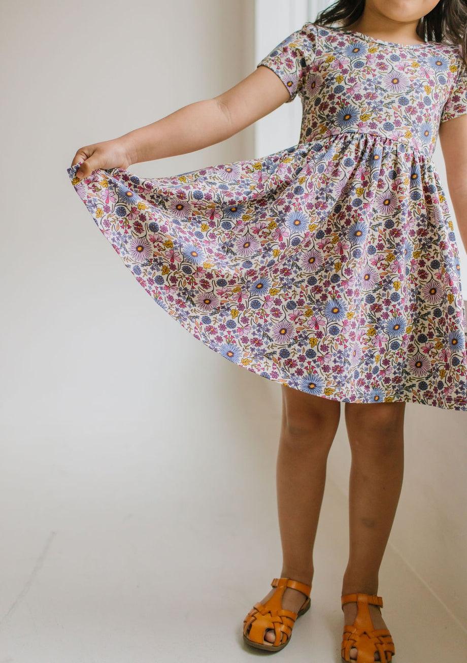 little girl wearing the purple floral dress, short sleeves with pops of blue, purple, pink and gold. she is holding the skirt out to show it is full and can fully twirl. 