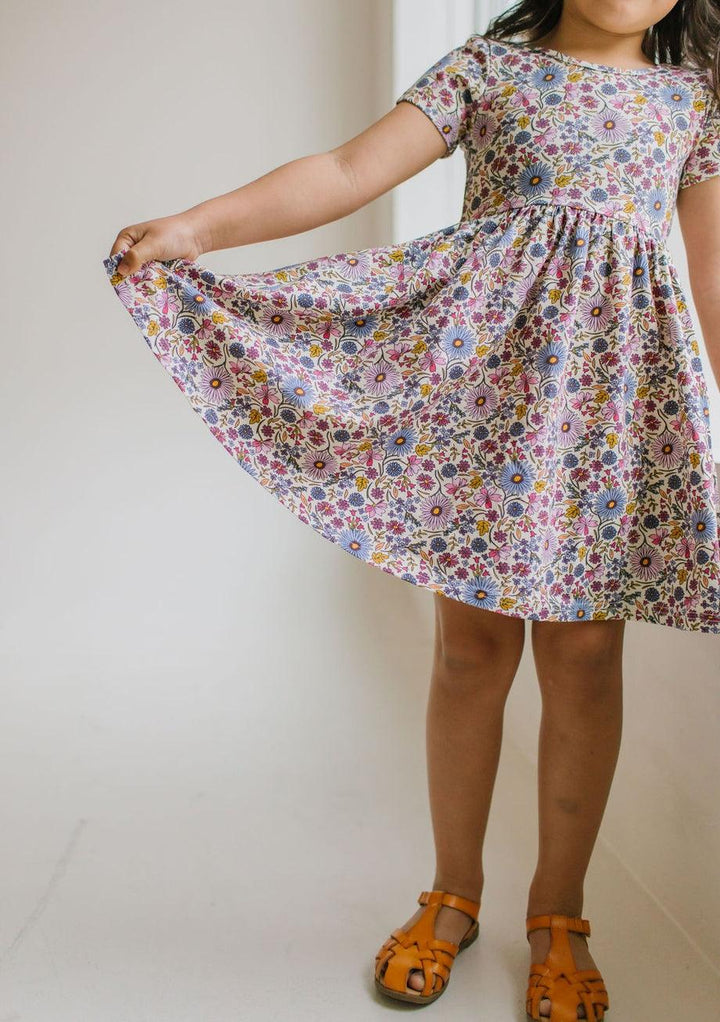 little girl wearing the purple floral dress, short sleeves with pops of blue, purple, pink and gold. she is holding the skirt out to show it is full and can fully twirl. 