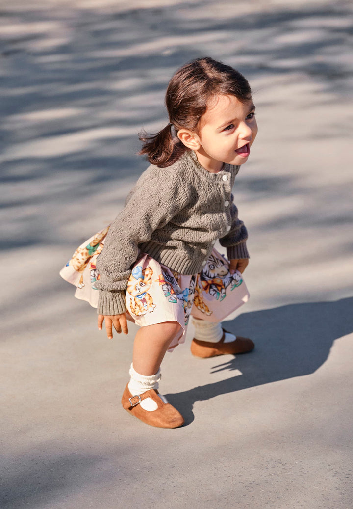 Child in a gray sweater and patterned dress walking on a concrete surface.