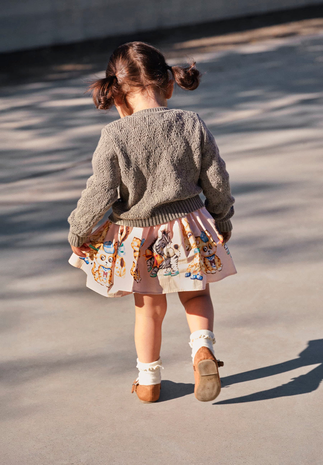 Child wearing a gray sweater and patterned skirt walking on a concrete surface.