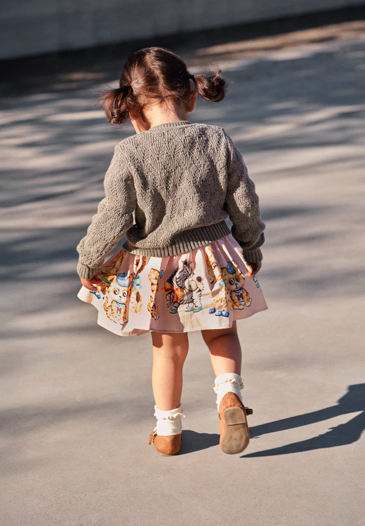 Child wearing a gray sweater and patterned skirt walking on a concrete surface.