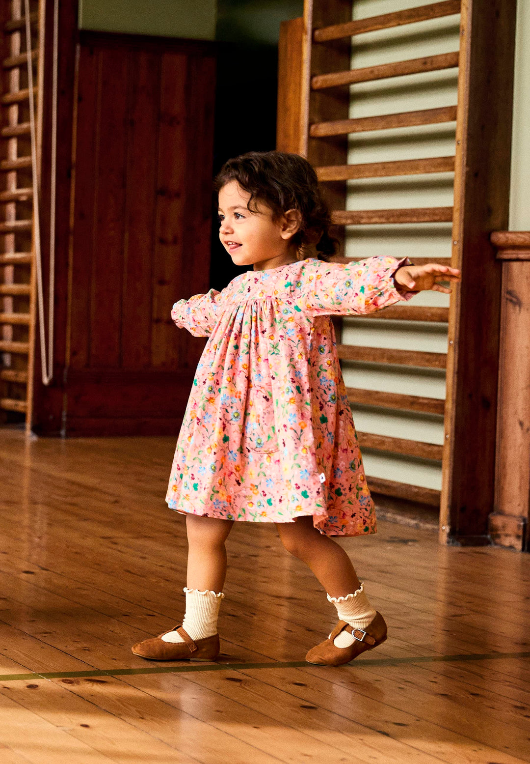 Young girl in a floral dress standing on a wooden floor with a staircase in the background.