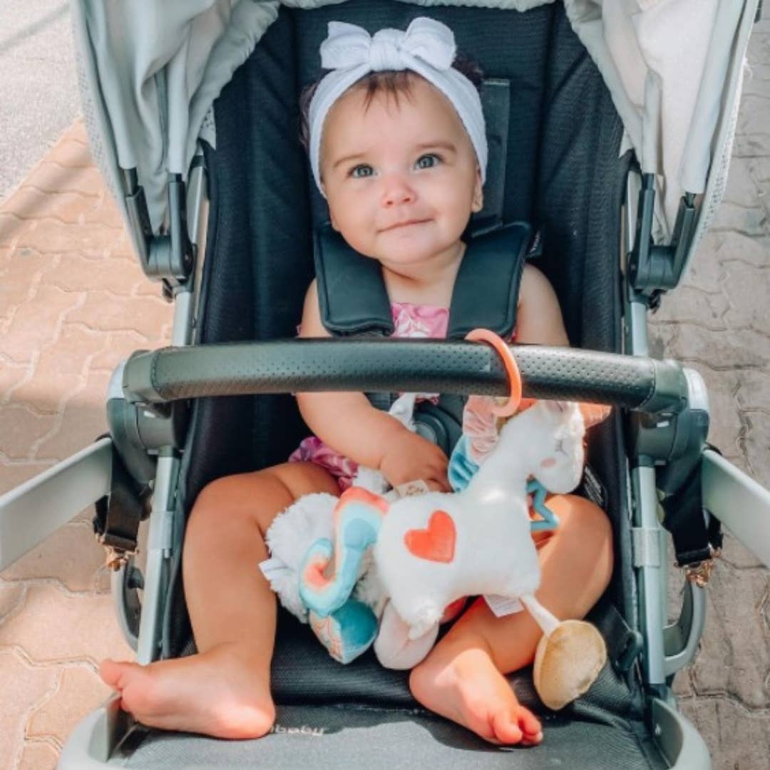 Baby sitting in a stroller with a toy, wearing a headband and holding a plush toy.