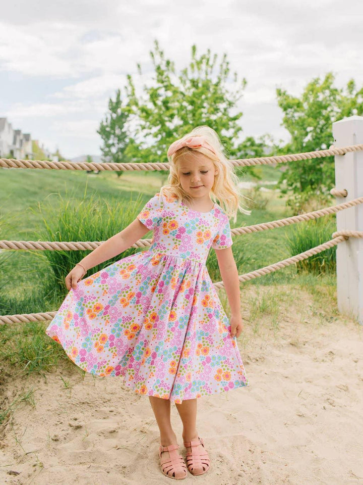 young girl wearing a multi colored floral dress, short sleeves, knee length. vibrant colored flowers: pink, blue, orange and purple. 