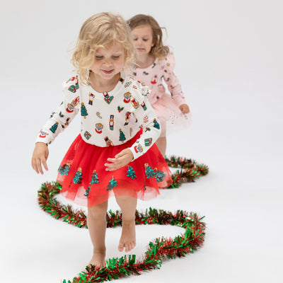 2 girls modeling the holiday sets that have holiday cookies on the shirt and a tutu skirt with sequins on it. the girl in the front has a white top with holiday cookies on it and a red tutu with sequin trees on it. 