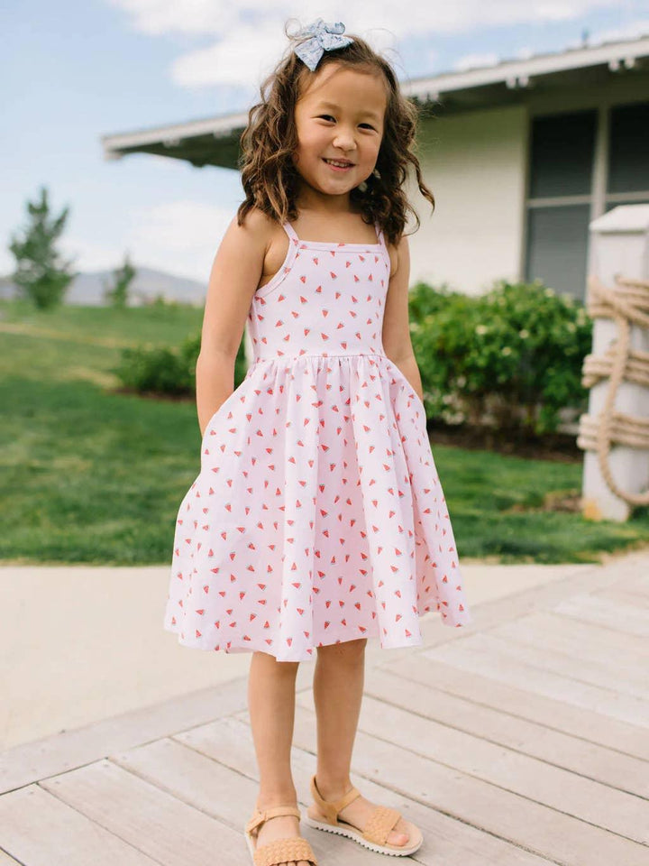 little girl wearing a summer dress, spaghetti strap, knee length. light pink and tiny watermelon slices all over the dress. 