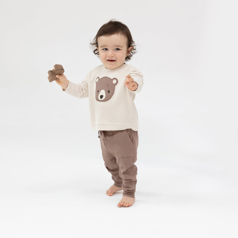 toddler modeling a thermal waffle outfit, ivory sweatshirt with teddy bear face on it and brown joggers with drawstring.