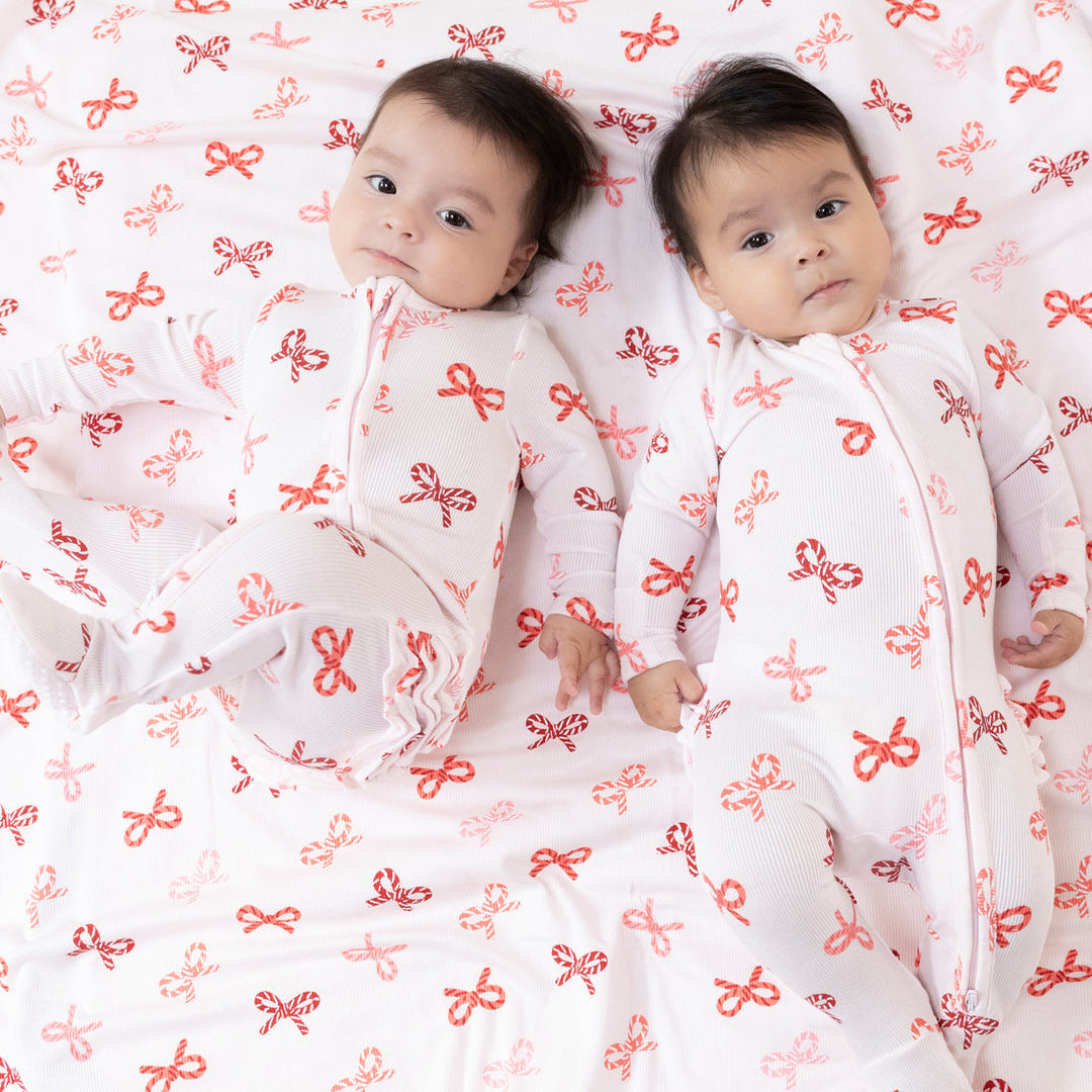Two babies in white onesies with red candy cane bows patterns lying on a white surface.