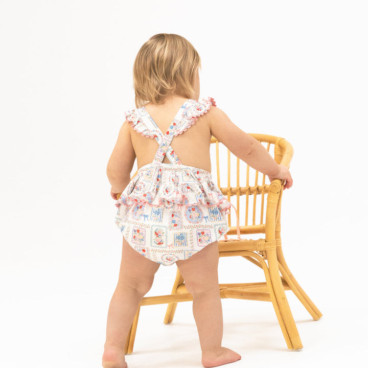 Child wearing a floral romper sitting on a wooden chair against a white background