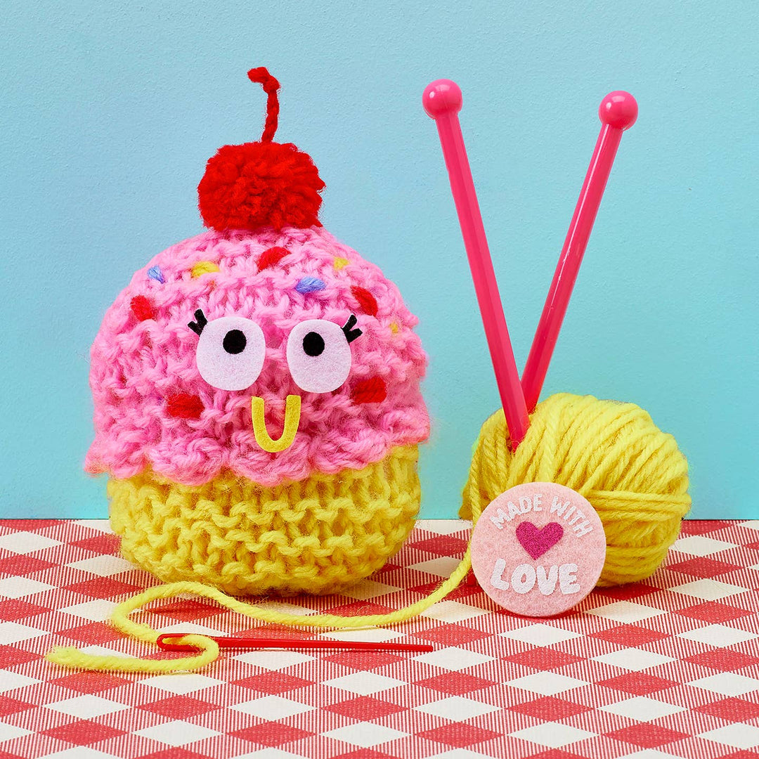 Crocheted cupcake character with pink frosting and red cherry on top, next to knitting needles and a ball of yellow yarn on a checkered tablecloth.