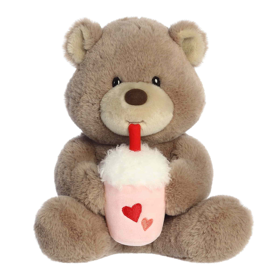 Plush teddy bear holding a pink cup with a red straw and hearts on a white background