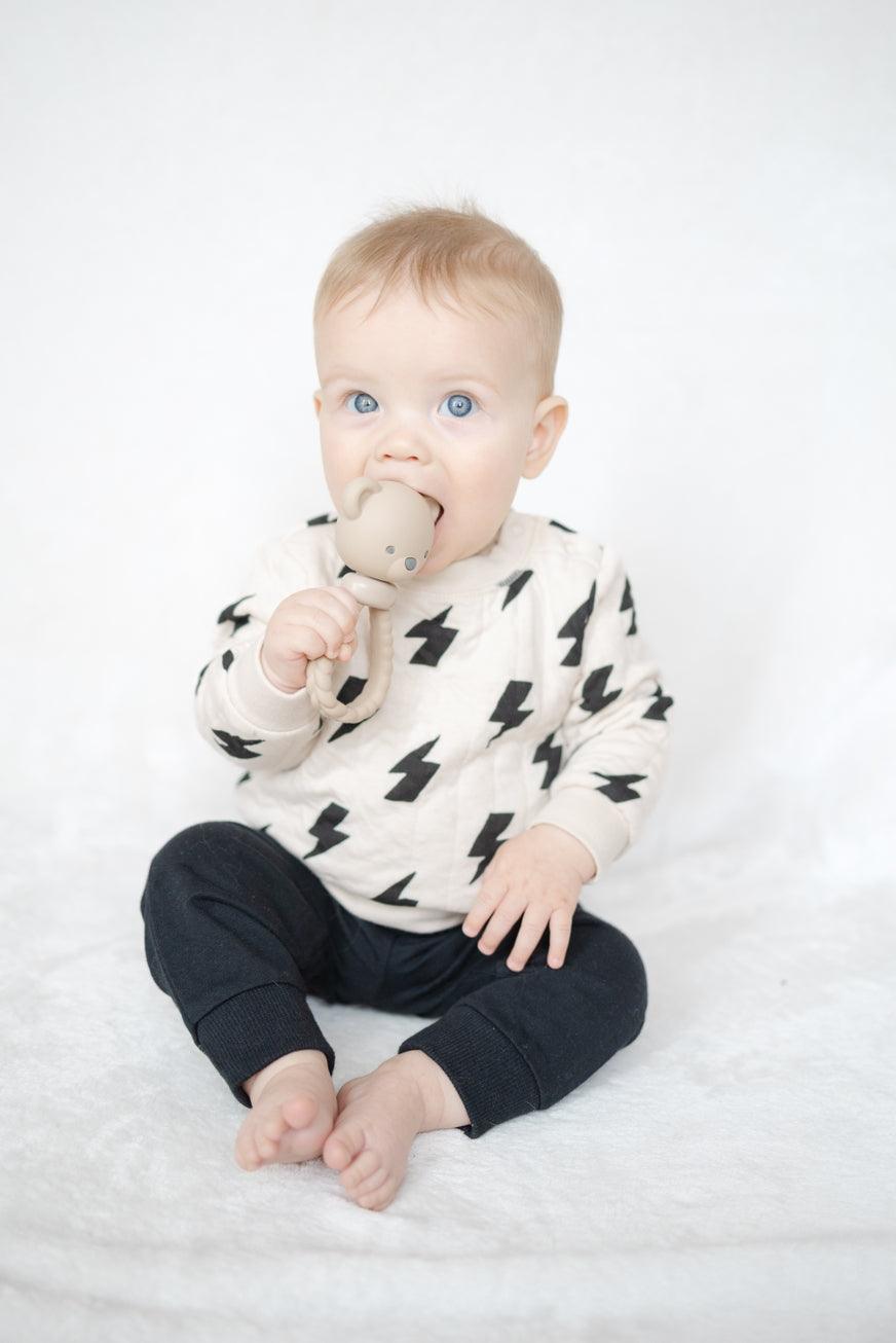 baby holding onto the teddy bear rattle and chewing on the head. 