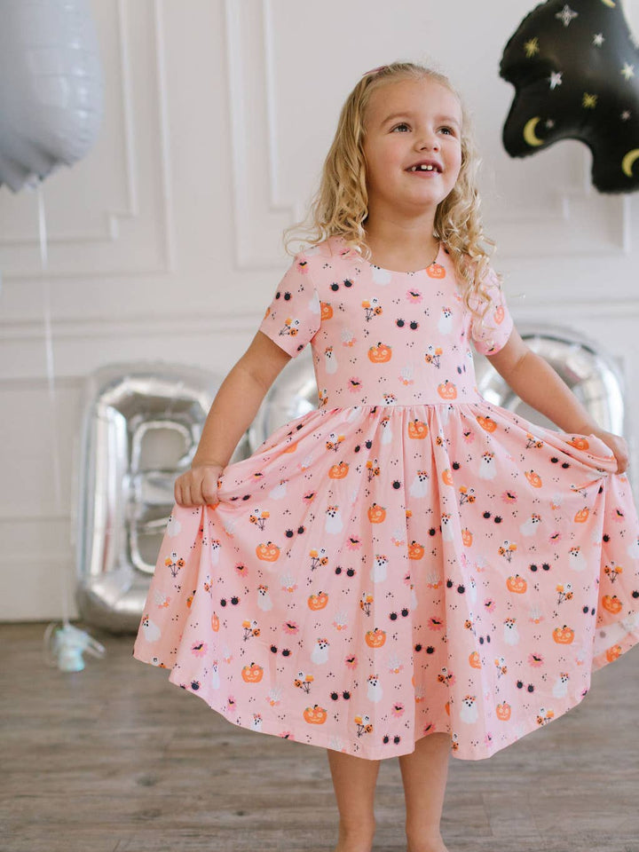 Young girl in a pink dress with Halloween patterns, standing indoors with balloons in the background.