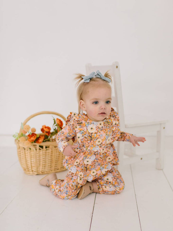 Baby in a floral outfit sitting on a white floor with a basket of flowers in the background