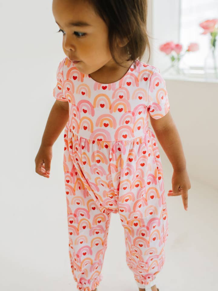 little baby modeling a jumpsuit with rainbows allover it and a heart in the center of the rainbow. 