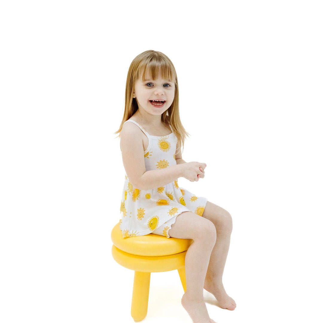little girl sitting on a yellow stool wearing the peplum tank and shorts with happy suns all over it. 