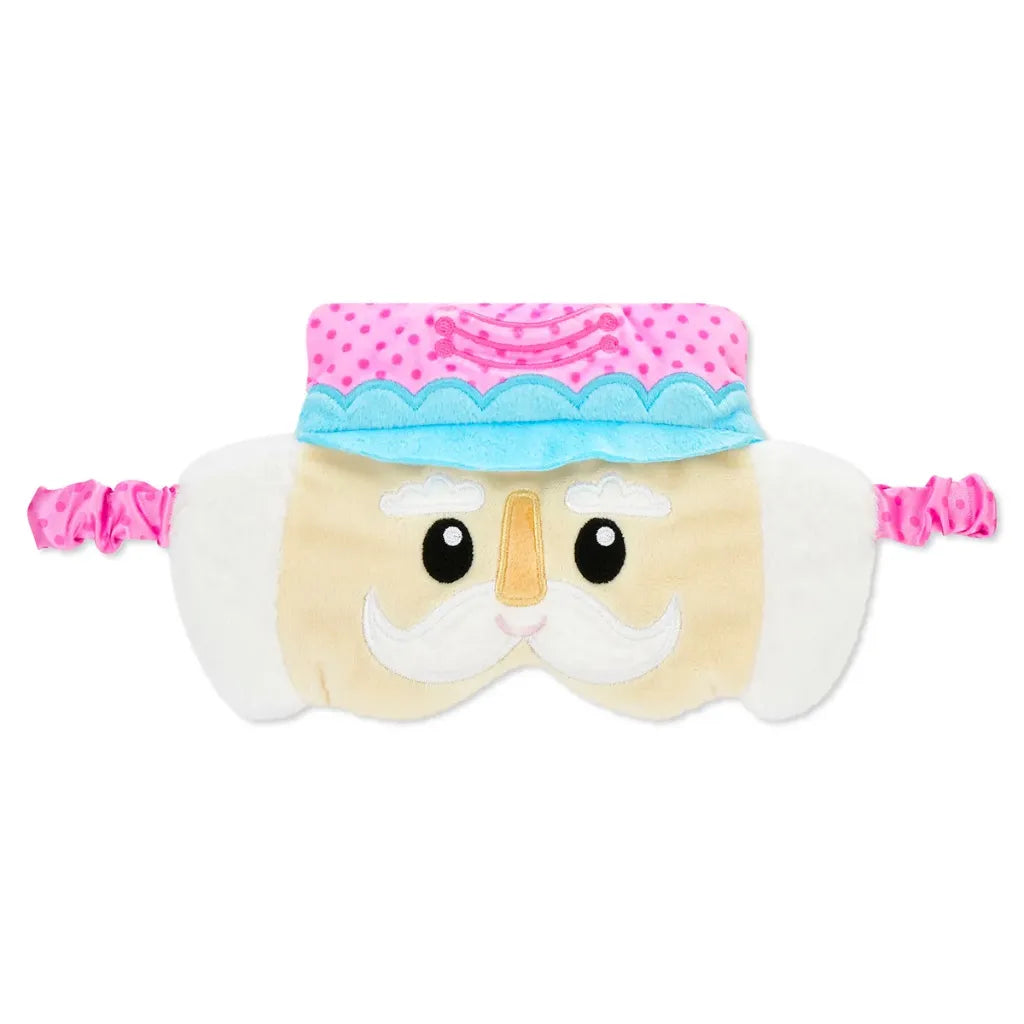 A plush sleep mask shaped like a nutcracker face, featuring a cream-colored body, white embroidered mustache, black eyes, a light blue and pink hat, and pink elastic straps on both sides.