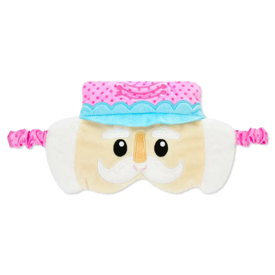 A plush sleep mask shaped like a nutcracker face, featuring a cream-colored body, white embroidered mustache, black eyes, a light blue and pink hat, and pink elastic straps on both sides.