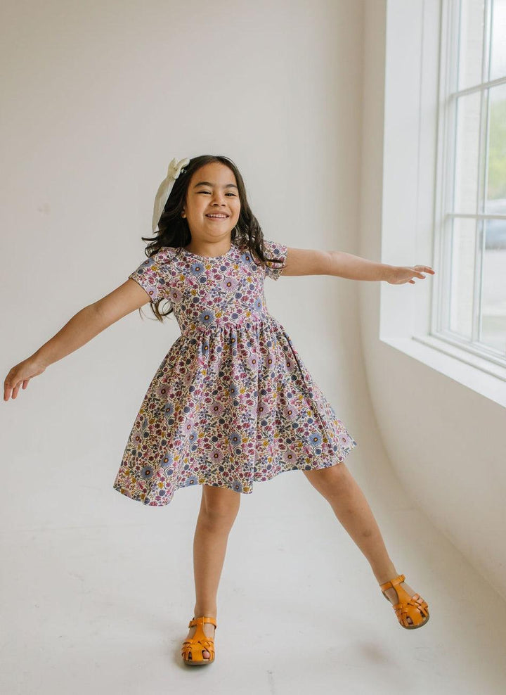 little girl modeling the short sleeve floral dress with flowers that are lavender, purple, blue, light pink and gold