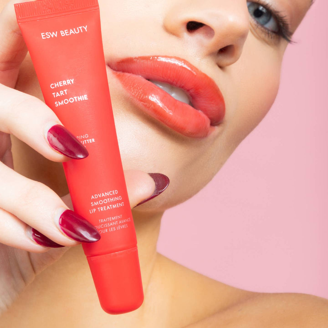 Red lip balm tube labeled 'ESW Beauty' held by a hand with red nail polish, against a pink background.