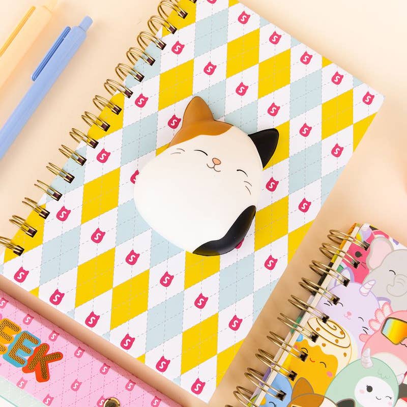 Notebook with squishmallow cat design on a checkered pattern, surrounded by other notebooks.