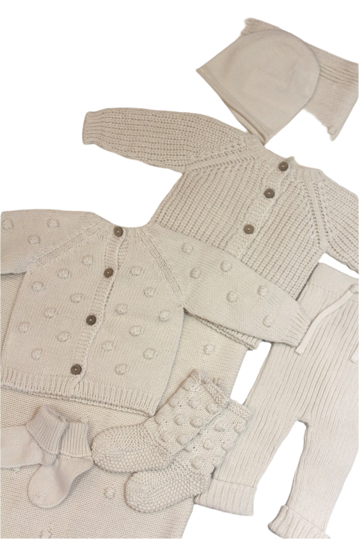 picture of creme colored pieces photographed together, laid out flat: two different sweaters, knit pants, socks and booties. all knits for baby