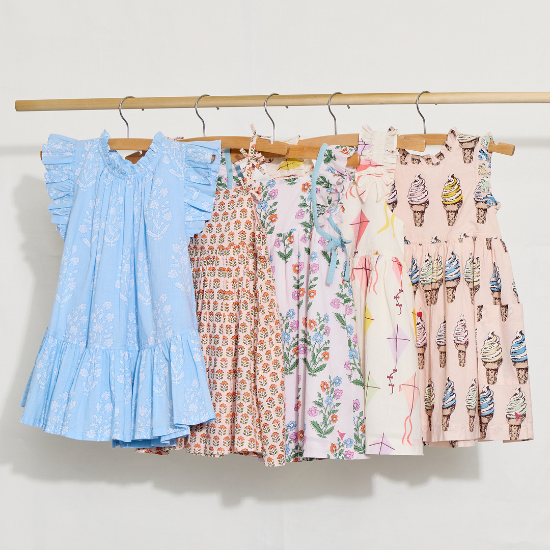 Five children's dresses with different patterns hanging on a rack against a white background
